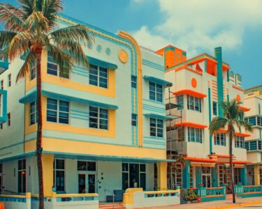 Palm trees line pastel Art Deco buildings along a sunny street under a bright blue sky with cream and coral facades.