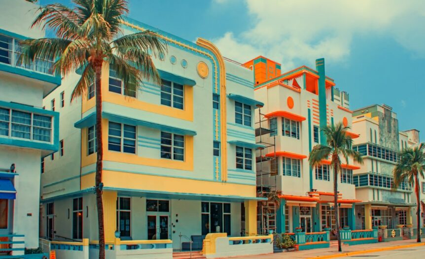 Palm trees line pastel Art Deco buildings along a sunny street under a bright blue sky with cream and coral facades.