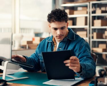 A man working at a laptop beside printed documents and cardboard boxes in a bright office workspace.