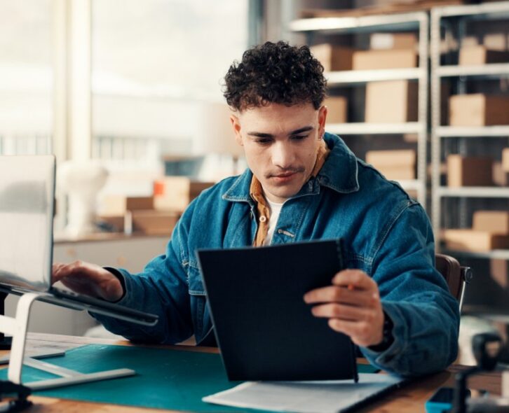 A man working at a laptop beside printed documents and cardboard boxes in a bright office workspace.