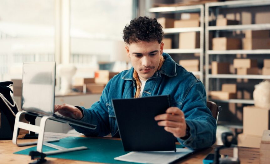 A man working at a laptop beside printed documents and cardboard boxes in a bright office workspace.