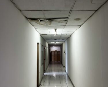 A poorly lit hallway with dilapidated ceiling tiles. The tiles are water stained and falling down into the hallway.