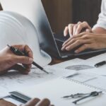 A close-up of two people sitting at a table with construction plans and diagrams, with a hard hat and a laptop.