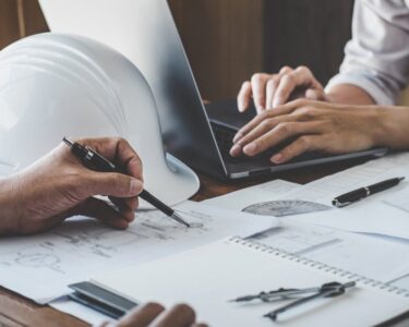 A close-up of two people sitting at a table with construction plans and diagrams, with a hard hat and a laptop.