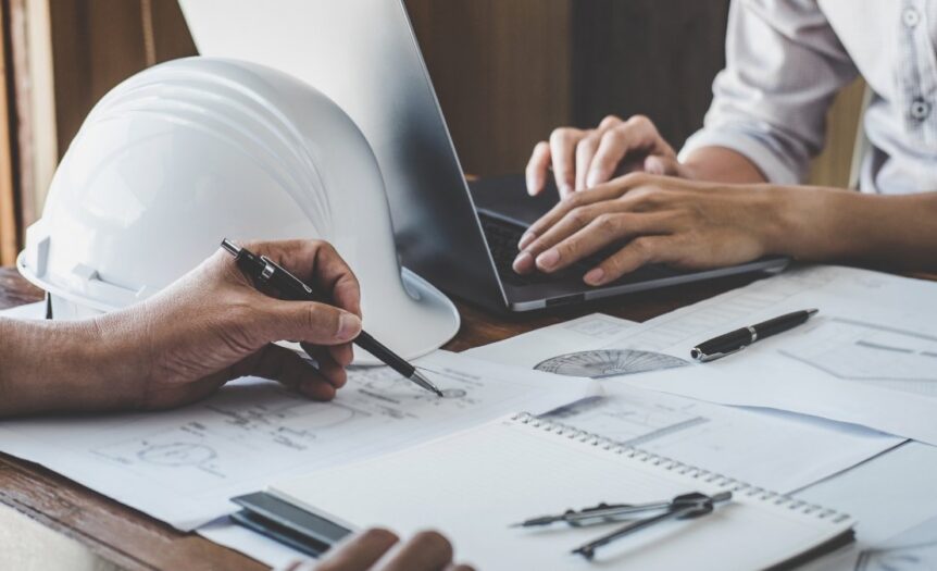 A close-up of two people sitting at a table with construction plans and diagrams, with a hard hat and a laptop.