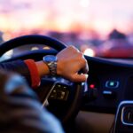 A person holding a steering wheel inside a car at dusk, with dashboard controls and blurred city lights ahead.