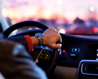 A person holding a steering wheel inside a car at dusk, with dashboard controls and blurred city lights ahead.