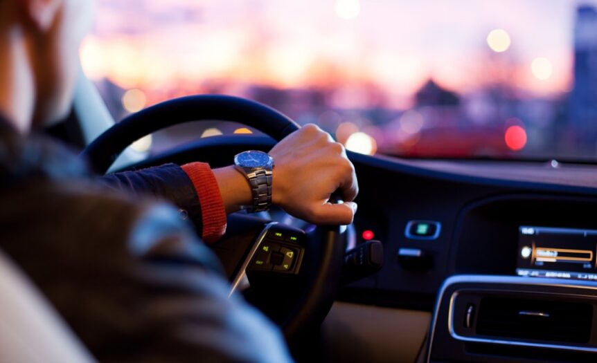 A person holding a steering wheel inside a car at dusk, with dashboard controls and blurred city lights ahead.