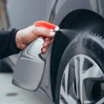 A person is kneeling down next to a car. They are using a spray bottle to spray the tire with water.