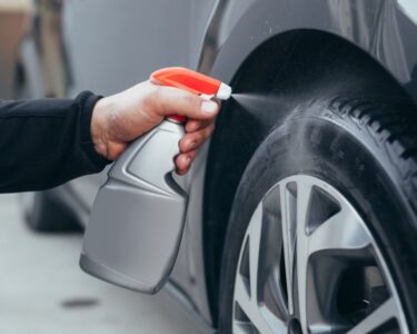 A person is kneeling down next to a car. They are using a spray bottle to spray the tire with water.