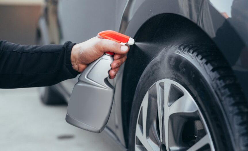 A person is kneeling down next to a car. They are using a spray bottle to spray the tire with water.