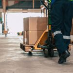 A person in a high-vis vest is using a jack to push a pallet filled with cardboard boxes through a warehouse.