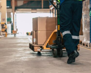 A person in a high-vis vest is using a jack to push a pallet filled with cardboard boxes through a warehouse.