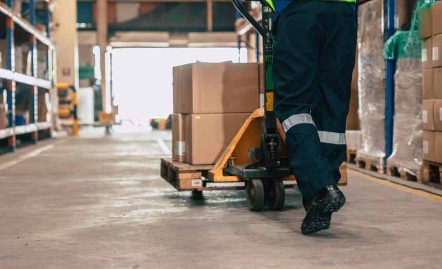 A person in a high-vis vest is using a jack to push a pallet filled with cardboard boxes through a warehouse.