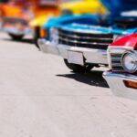 A red classic car leads a bright row of vintage cars with open hoods at a sunny outdoor Miami car show.