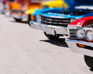 A red classic car leads a bright row of vintage cars with open hoods at a sunny outdoor Miami car show.