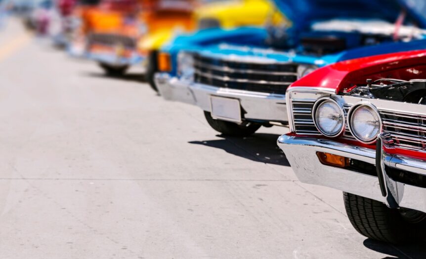A red classic car leads a bright row of vintage cars with open hoods at a sunny outdoor Miami car show.