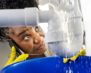 A woman leaning under a sink and holding a bucket under a leaking pipe. She has on yellow gloves and is on the phone.