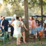 A large gathering of people are outside in an area with several trees. The guests are blurry in the background.