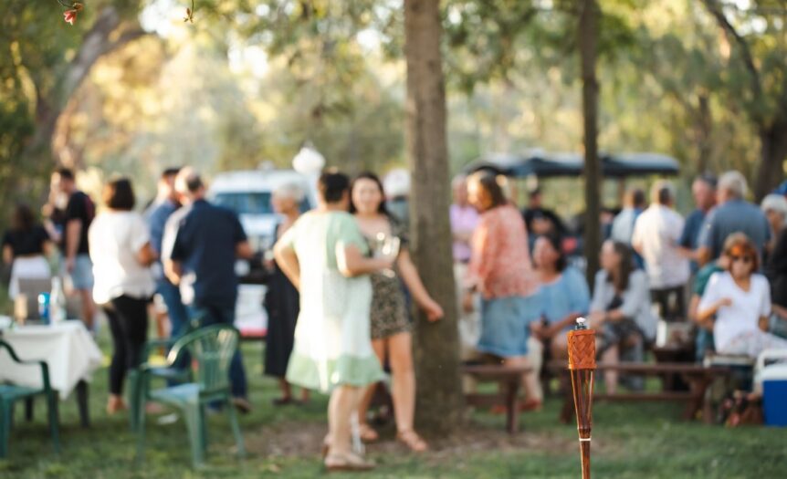 A large gathering of people are outside in an area with several trees. The guests are blurry in the background.