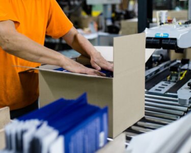 A woman in an orange T-shirt packing a cardboard box on a conveyor belt at the end of a packaging line.