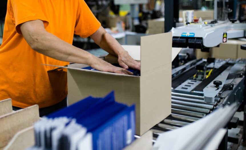 A woman in an orange T-shirt packing a cardboard box on a conveyor belt at the end of a packaging line.
