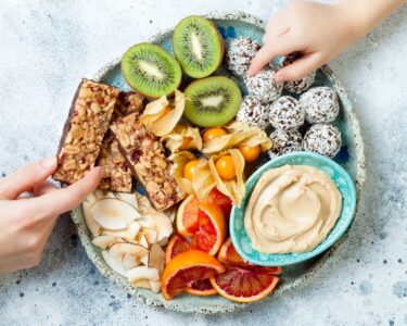 Colorful snack platter holds sliced kiwi, citrus, coconut bites, nut bars, berries, coconut chips, and creamy dip.