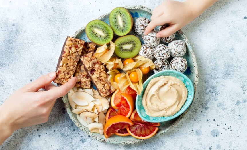 Colorful snack platter holds sliced kiwi, citrus, coconut bites, nut bars, berries, coconut chips, and creamy dip.