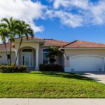 A one-story home with orange, clay roof tiles, a concrete driveway, green grass in the front yard, and three palm trees.