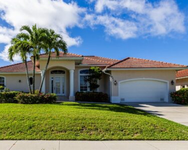 A one-story home with orange, clay roof tiles, a concrete driveway, green grass in the front yard, and three palm trees.