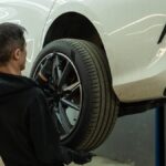 A mechanic wearing black gloves and replacing the rear driver-side tire on a white car in a repair shop.