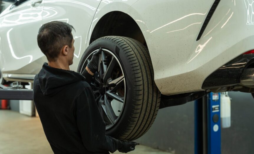 A mechanic wearing black gloves and replacing the rear driver-side tire on a white car in a repair shop.