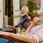 A smiling older couple relaxes in outdoor chairs with cold drinks beside a recreational vehicle on a sunny day.