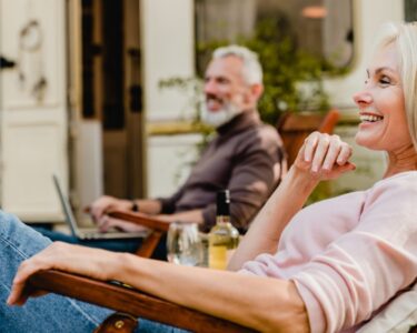 A smiling older couple relaxes in outdoor chairs with cold drinks beside a recreational vehicle on a sunny day.