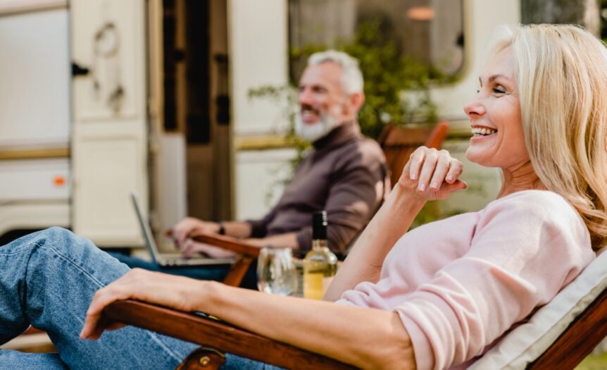 A smiling older couple relaxes in outdoor chairs with cold drinks beside a recreational vehicle on a sunny day.