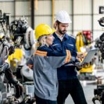 Two manufacturing workers in hard hats inspect industrial machinery while reviewing information on a laptop.