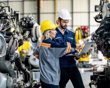Two manufacturing workers in hard hats inspect industrial machinery while reviewing information on a laptop.