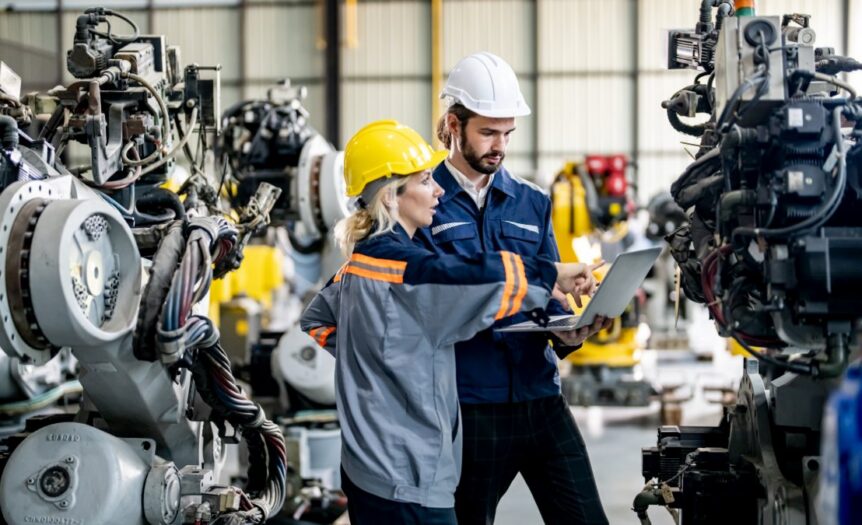 Two manufacturing workers in hard hats inspect industrial machinery while reviewing information on a laptop.