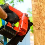 Construction worker wearing black gloves uses a red pneumatic nail gun to secure plywood sheathing to a wooden frame.