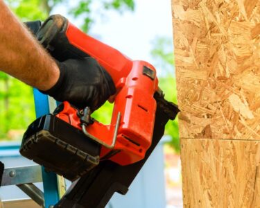 Construction worker wearing black gloves uses a red pneumatic nail gun to secure plywood sheathing to a wooden frame.