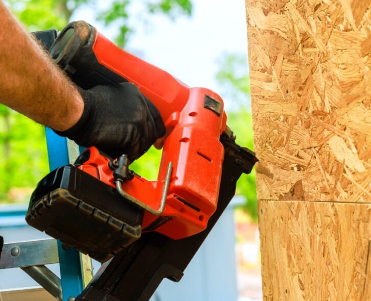 Construction worker wearing black gloves uses a red pneumatic nail gun to secure plywood sheathing to a wooden frame.