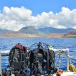 Scuba gear sits on a bench next to a gas cylinder on a boat. The boat is floating on water near a tropical island.