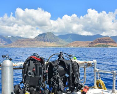 Scuba gear sits on a bench next to a gas cylinder on a boat. The boat is floating on water near a tropical island.