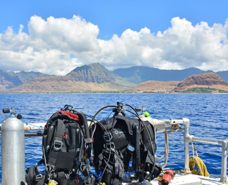 Scuba gear sits on a bench next to a gas cylinder on a boat. The boat is floating on water near a tropical island.