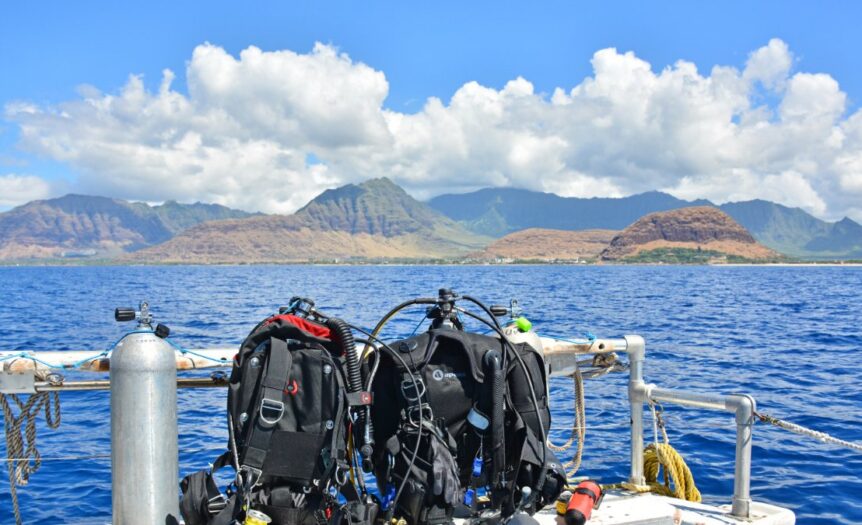Scuba gear sits on a bench next to a gas cylinder on a boat. The boat is floating on water near a tropical island.